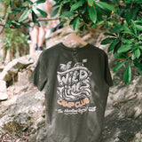A khaki colored t-shirt with text and graphic design, displayed in a natural outdoor setting with trees and a person in the background.