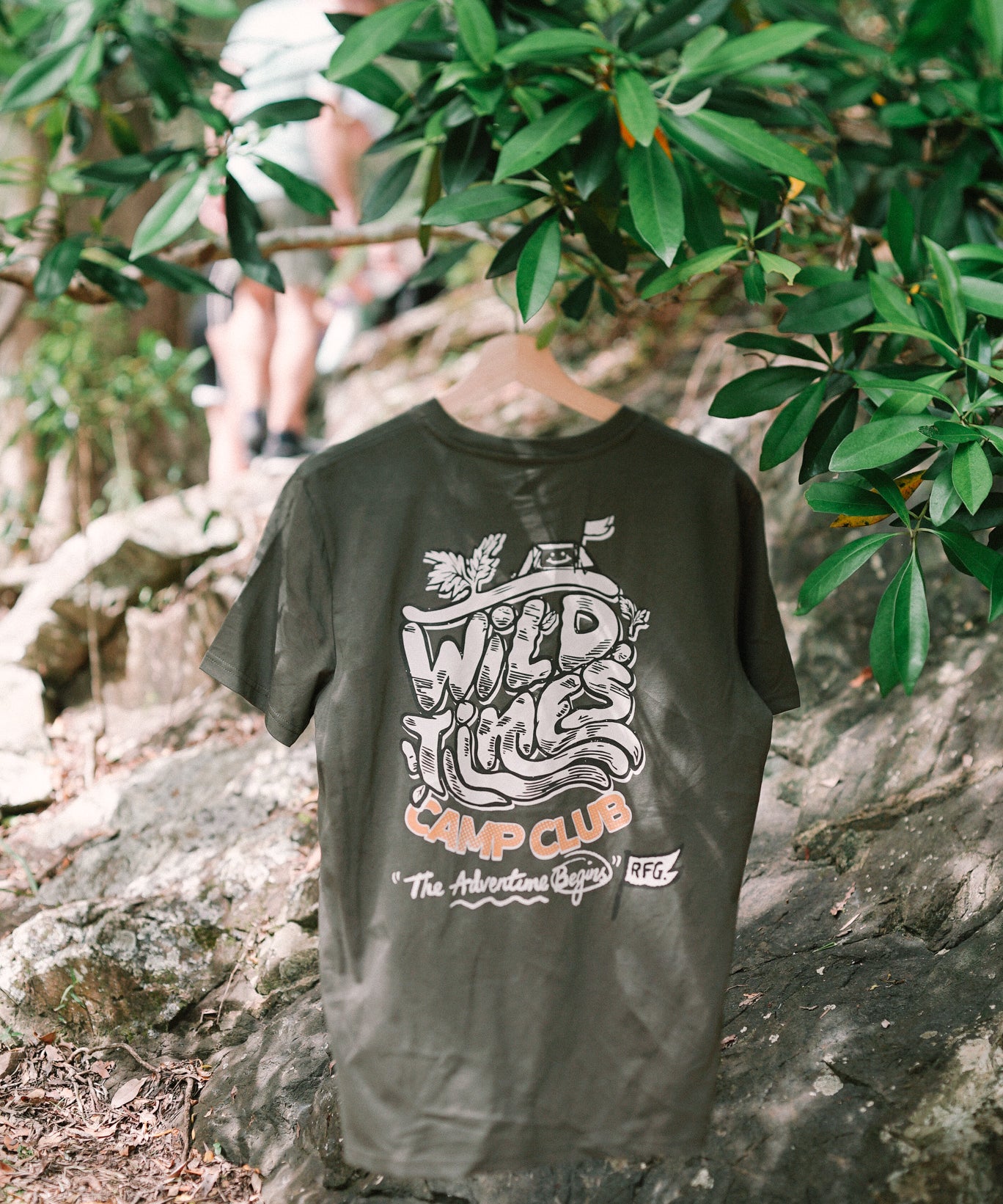A khaki colored t-shirt with text and graphic design, displayed in a natural outdoor setting with trees and a person in the background.
