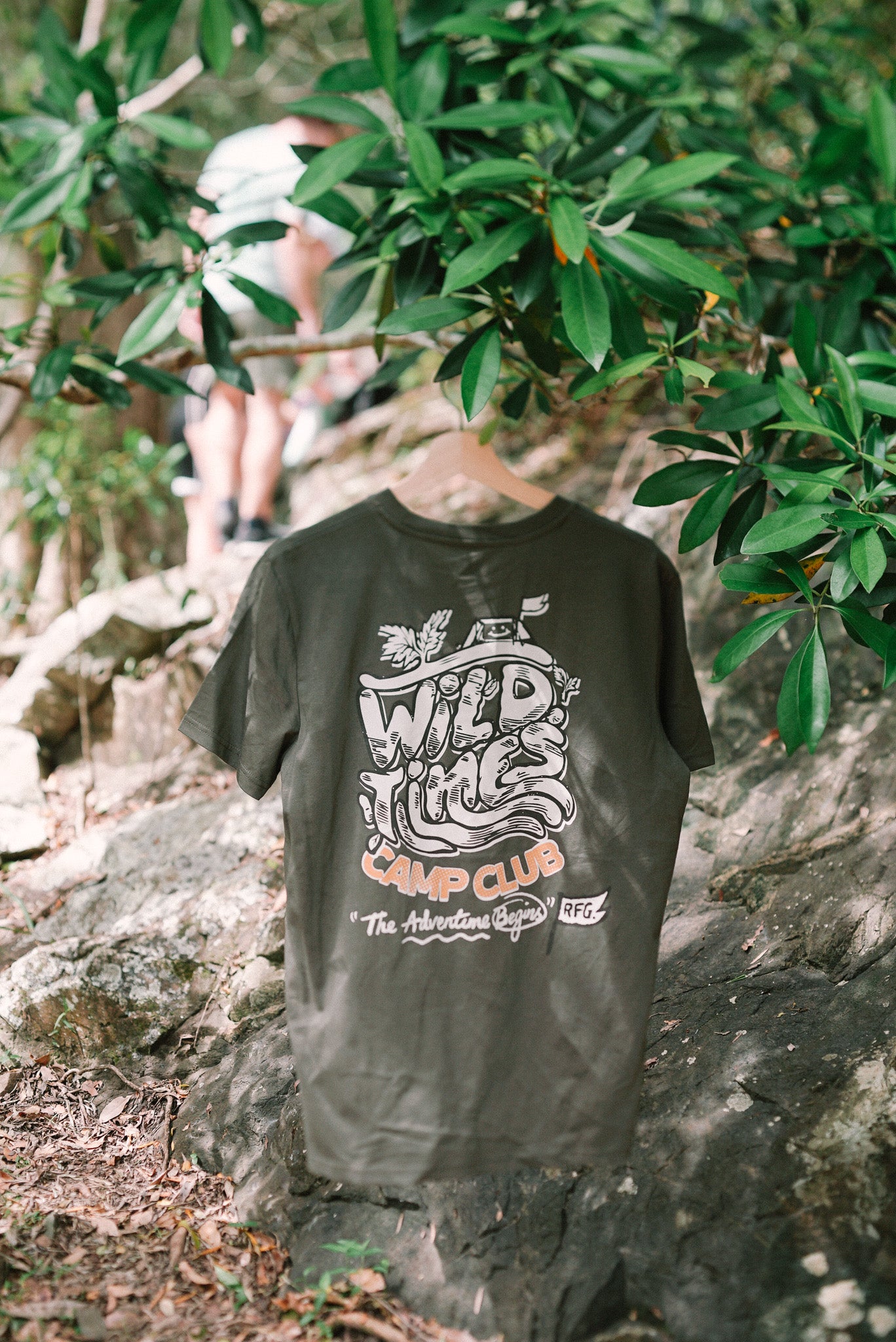 A khaki colored t-shirt with text and graphic design, displayed in a natural outdoor setting with trees and a person in the background.